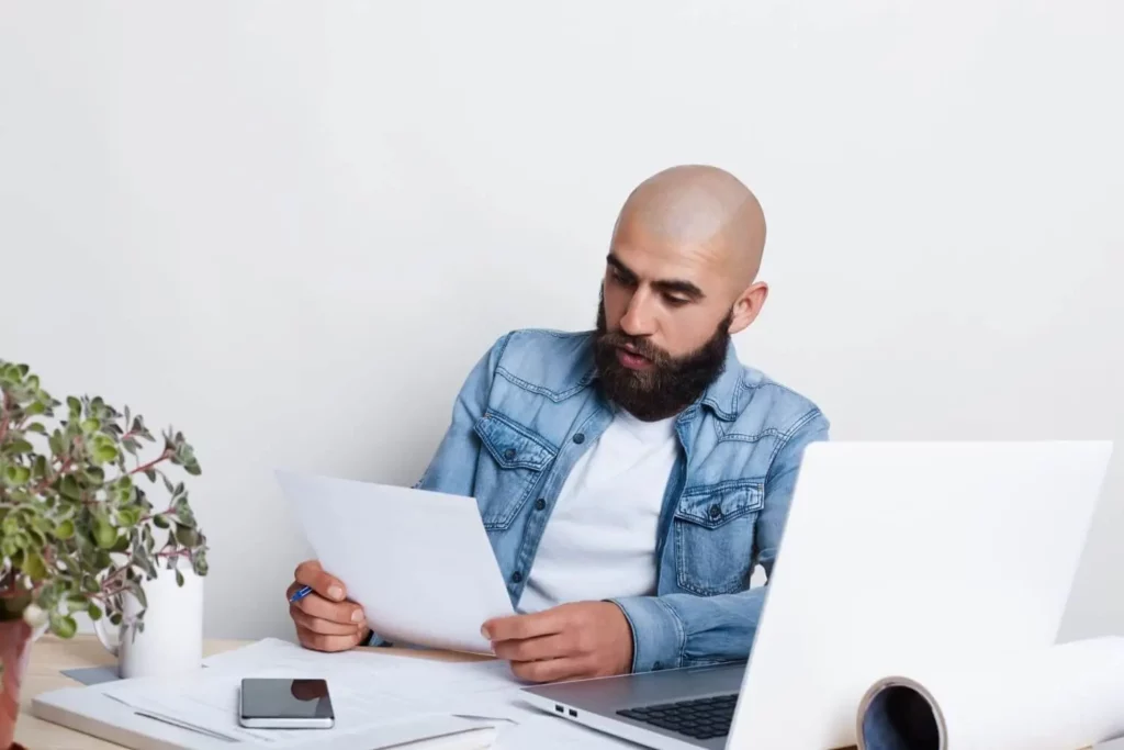 Um homem barbudo está sentado à mesa com um laptop, segurando papéis e uma caneta, parecendo concentrado. Há uma planta, um pedaço de papel enrolado e um smartphone sobre a mesa. O fundo é branco liso.