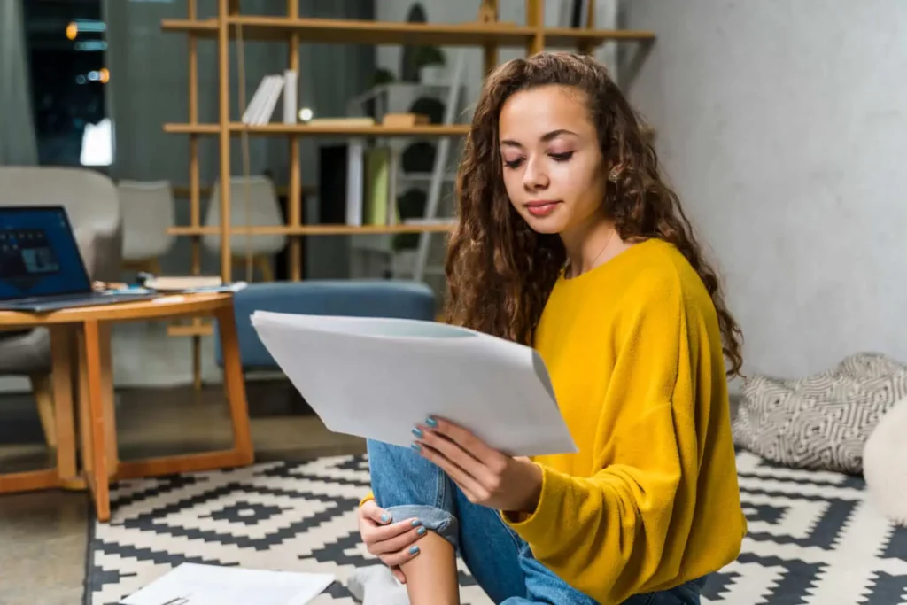Imagem de uma jovem com cabelos longos e cacheados, vestindo um suéter amarelo e jeans, sentada em um tapete estampado enquanto lê jornais. Um notebooke prateleiras com livros aparecem ao fundo, conferindo ao ambiente um ar aconchegante e moderno.