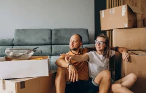 Imagem de uma mulher sorridente e de um homem pensativo sentados no chão, em meio a caixas de mudança, na sala de estar. A mulher está apontando para a frente, e o homem parece pensativo. Um sofá e objetos ainda não desembalados podem ser vistos ao fundo.