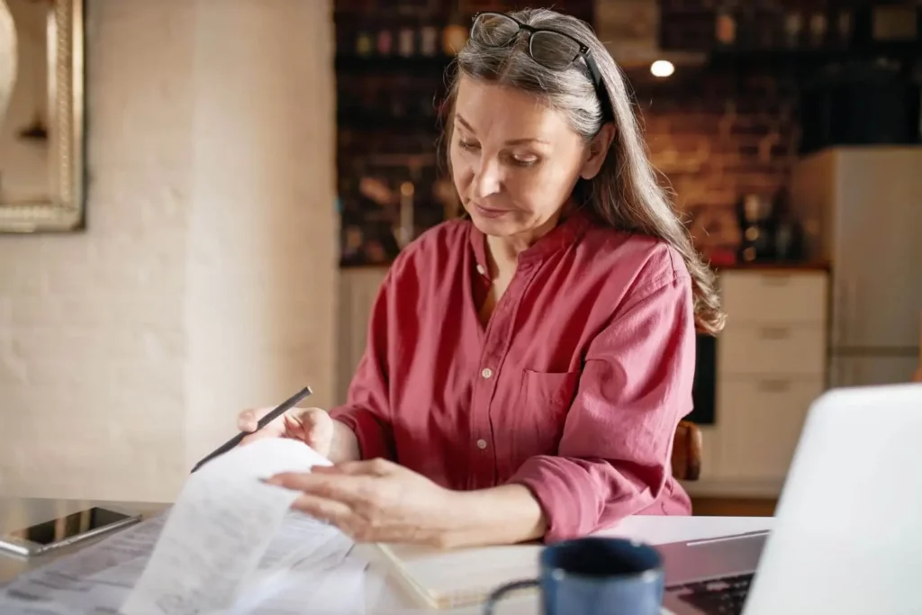 Imagem de uma mulher de óculos e camisa vermelha examinando alguns papéis em uma mesa, com um notebook, um caderno e uma caneca de café, em um ambiente aconchegante de cozinha.