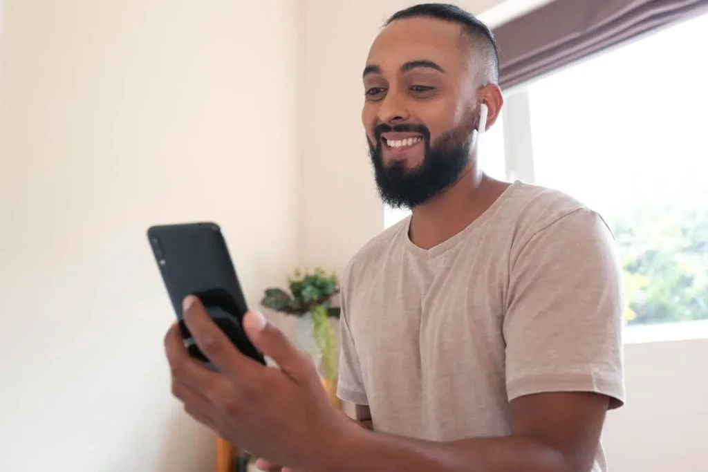 Imagem de um homem sorridente, de barba, vestindo uma camiseta clara e fones de ouvido sem fio, olhando para o smartphone em uma sala bem iluminada, com uma janela e uma planta ao fundo.