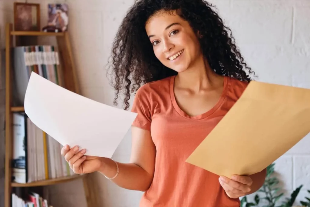 Imagem de uma jovem sorridente, de cabelos cacheados, segurando uma folha de papel em branco com uma das mãos e um grande envelope amarelo com a outra, em pé, em um ambiente com estantes de livros e uma parede branca ao fundo.