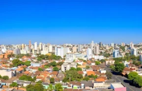 Vista panorâmica do horizonte de uma cidade, com prédios altos ao fundo e casas com telhados vermelhos e árvores verdes em primeiro plano, sob um céu azul límpido.