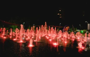 À noite, dezenas de fontes iluminadas em vermelho jorram água para cima em uma praça aberta, com prédios, árvores e pessoas visíveis ao fundo.