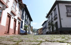Vista ao nível da rua de uma rua de paralelepípedos ladeada por prédios históricos em estilo colonial, alguns com varandas e plantas, sob um céu azul e límpido. Colinas verdes são visíveis ao fundo, à distância.