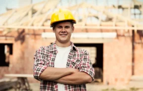 Imagem de um operário da construção civil, usando um capacete amarelo e uma camisa xadrez, em frente a um prédio em construção, sorrindo com os braços cruzados, para ilustrar matéria sobre construção em área de APP.