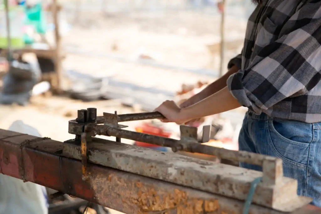 Imagem de uma pessoa vestindo camisa xadrez e jeans, dobrando uma barra de metal com uma dobradeira manual enferrujada ao ar livre, para ilustrar matéria sobre o que é construção em área de APP