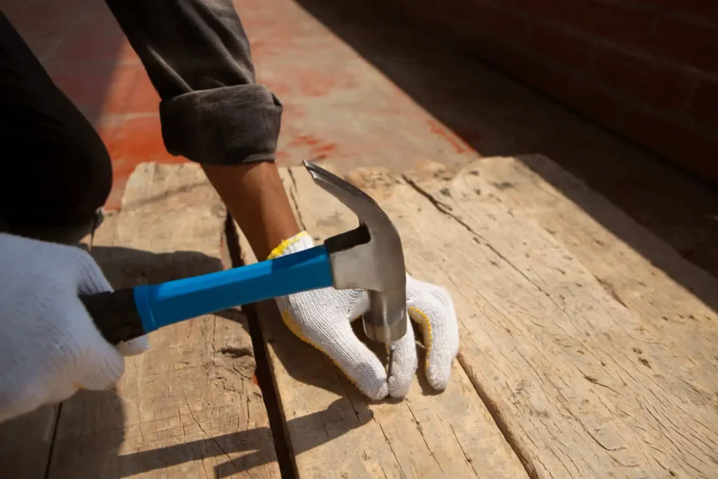Imagem de uma pessoa de luvas brancas usando um martelo de cabo azul para cravar um prego em uma tábua de madeira áspera, trabalhando ao ar livre sob a luz do sol, para ilustrar matéria sobre quando pode fazer uma construção em área de APP.
