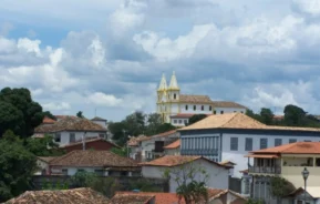 Uma cidade situada na encosta, com prédios de telhados de terracota e uma igreja branca imponente com duas torres, sob um céu nublado.
