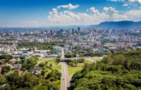 Vista aérea de uma cidade em expansão, com uma estrada em primeiro plano que leva a um horizonte repleto de arranha-céus e montanhas distantes, sob um céu azul.