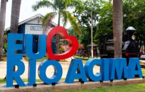 Um grande letreiro com os dizeres EU AMO RIO ACIMA, com um coração vermelho no lugar da letra A, localizado próximo a uma locomotiva a vapor histórica e a um prédio com a placa RIO ACIMA.