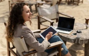 Imagem de uma mulher sentada em uma cadeira em uma praia, segurando um tablet e uma caneta digital, com um notebook aberto e uma xícara de café sobre uma mesa próxima para ilustrar matéria sobre comprar casa na praia com consórcio para alugar.