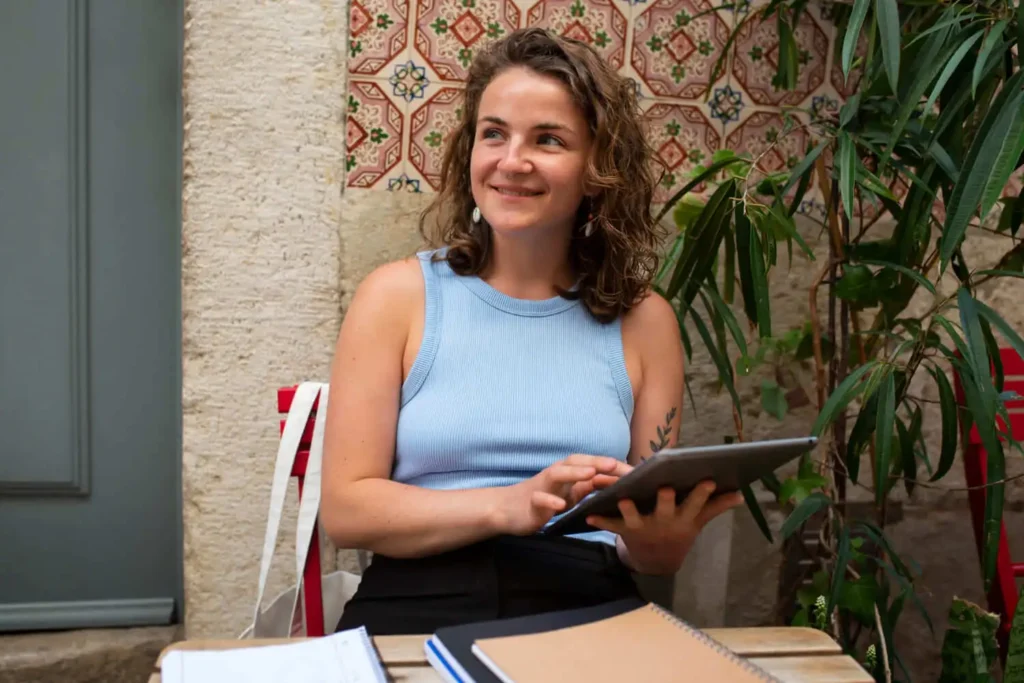 Imagem de uma jovem vestindo uma regata azul-clara, sentada em uma mesa ao ar livre, sorrindo enquanto desvia o olhar do tablet que segura nas mãos.