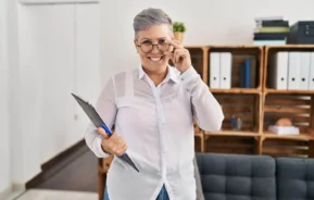 Imagem de uma mulher sorridente, de cabelo curto e grisalho, segurando uma prancheta e ajeitando os óculos em um escritório para ilustrar matéria sobre o consórcio para servidor público.